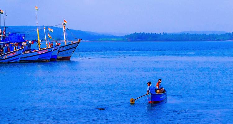 Dos hombres remando un pequeño bote azul en un mar tranquilo con barcos pesqueros anclados cerca.