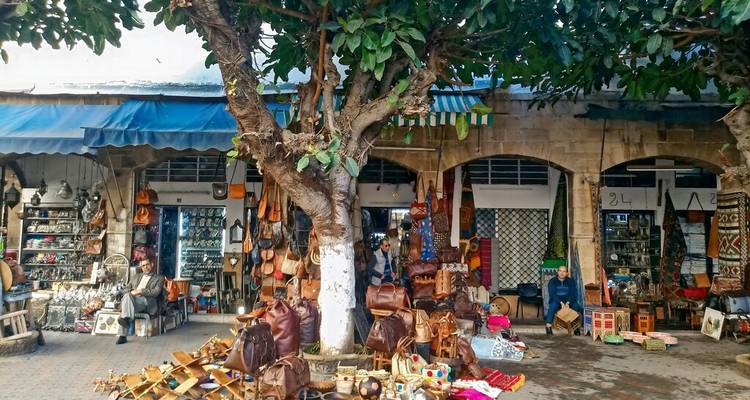 Marché extérieur animé avec des articles en cuir et de l'artisanat exposés sous un arbre ombragé.