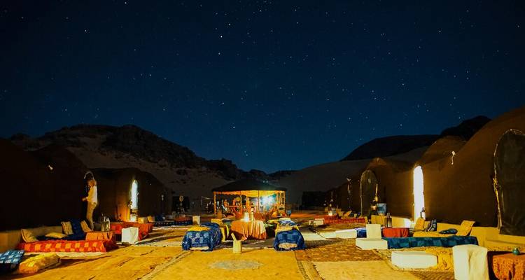 Site de glamping dans le désert étoilé avec des tapis colorés, des tables basses et des tentes qui brillent dans l'obscurité.
