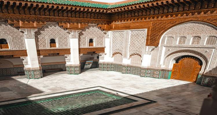 Cour ensoleillée d'une madrasa historique avec des stucs complexes, des carreaux de zellige et un petit bassin.