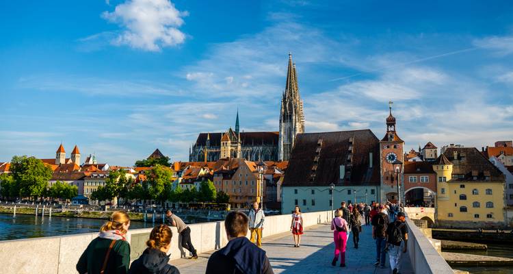 Les gens traversent l'historique Pont de Pierre vers les flèches de la cathédrale de Ratisbonne sous un ciel lumineux.