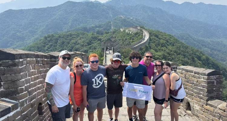 Lächelnde Reisegruppe posiert auf einem Abschnitt der Großen Mauer mit grünen Bergen, die sich dahinter erstrecken.