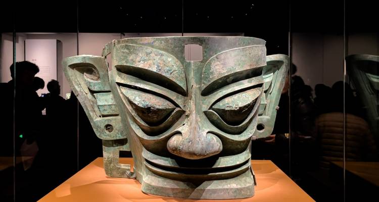 Große antike Bronzemaske, die in einem schwach beleuchteten Museum ausgestellt ist, mit Silhouetten von Besuchern im Hintergrund.