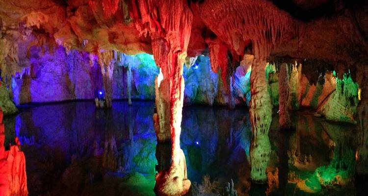 Farbig beleuchtete Stalaktiten und Stalagmiten, die sich in stillem Wasser in einer beleuchteten Höhle spiegeln.