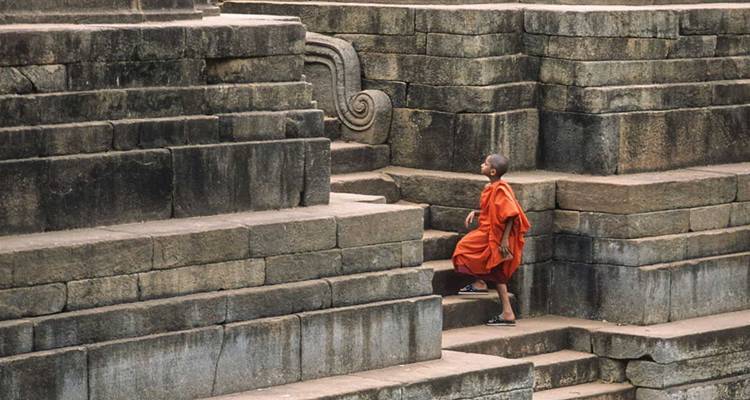 Buddhist monk in orange robes ascending ancient stone steps of a ruined temple complex