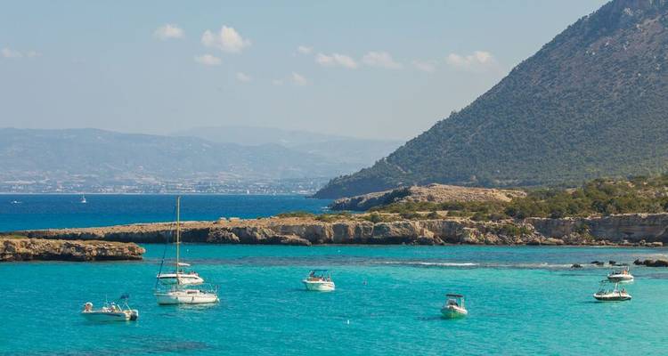 Turquoise bay with several anchored sailboats set against a wooded peninsula and distant mountains