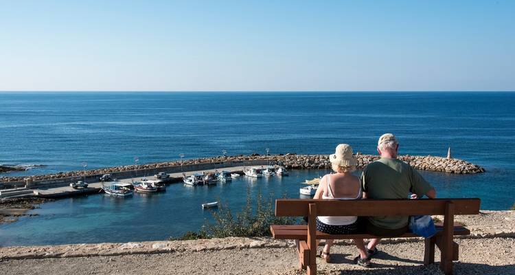 Senior couple sitting on a bench overlooking a small fishing harbour and open sea