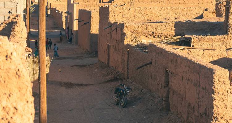 Calle estrecha y polvorienta del pueblo con muros de adobe, niños jugando y una motocicleta estacionada
