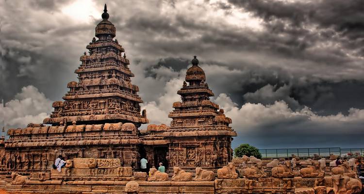 Ancient Shore Temple with dramatic dark clouds overhead and visitors walking around the intricate stone carvings.