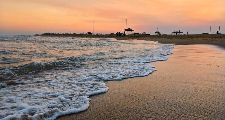 Gentle waves washing onto a sandy beach at sunset with silhouettes of umbrellas and people in the distance.