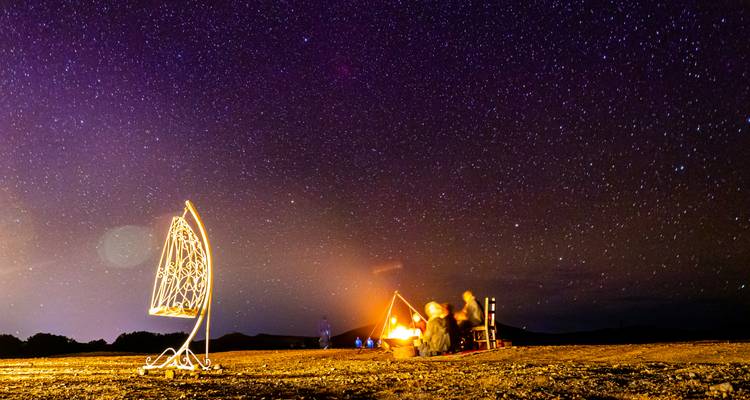Fogata del desierto nocturno bajo un cielo brillante lleno de estrellas con viajeros reunidos alrededor de las llamas.