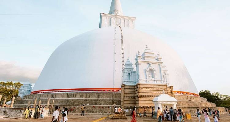 Eine massive weiße Stupa mit kleinen Besuchergruppen, die unter klarem blauem Himmel um deren Basis herumgehen.