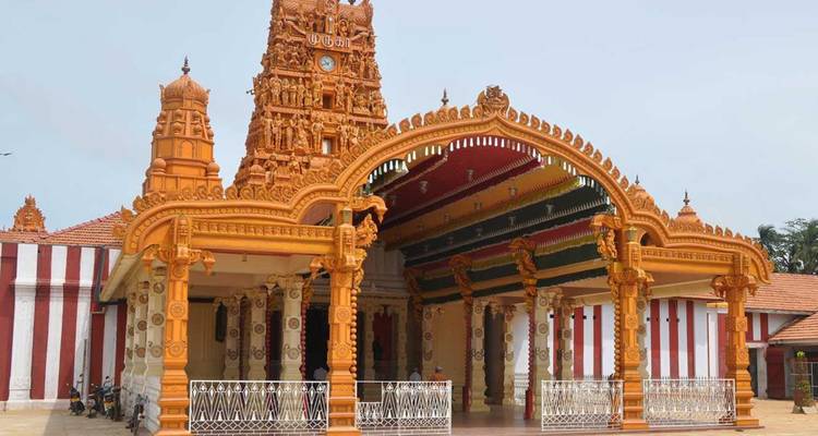 Ein verziertes orange-goldenes Hindu-Tempel-Tor mit detaillierten Schnitzereien und gestreiften Außenwänden vor einem blassen Himmel.