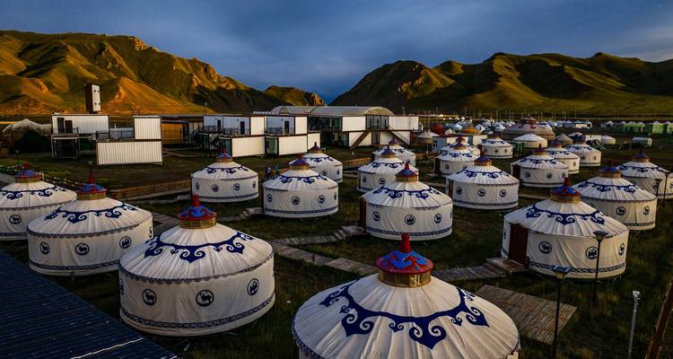 Des rangées de yourtes circulaires aux motifs bleus parsèment une plaine herbeuse sous des montagnes doucement éclairées au crépuscule.