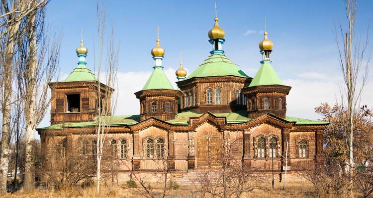 Une église orthodoxe rustique en bois aux toits verts et aux dômes en oignon dorés se dresse sous un ciel bleu clair.