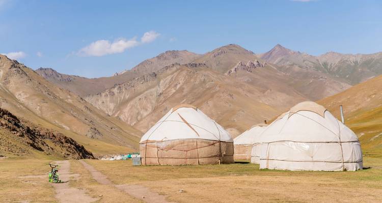 Trois yourtes blanches sont situées sur un plateau d'altitude herbeux entouré de montagnes arides sous un ciel dégagé.