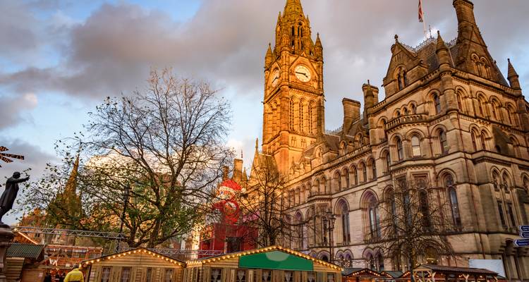 Der Uhrenturm des Rathauses von Manchester erhebt sich über festliche Marktstände unter dramatischen Abendwolken.