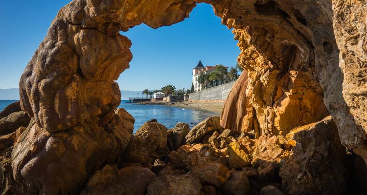 Une arche rocheuse encadre un manoir en bord de mer et une eau bleue calme par un matin clair.