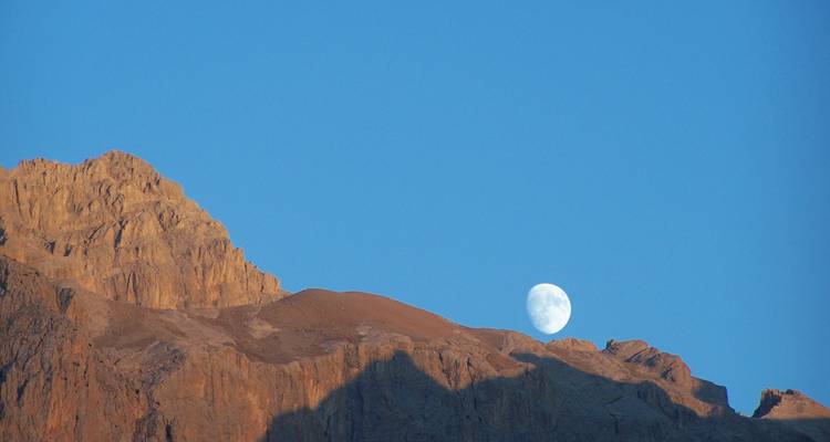 La pleine lune se couche derrière des pics rougeâtres escarpés sur un ciel d'aube bleu profond.