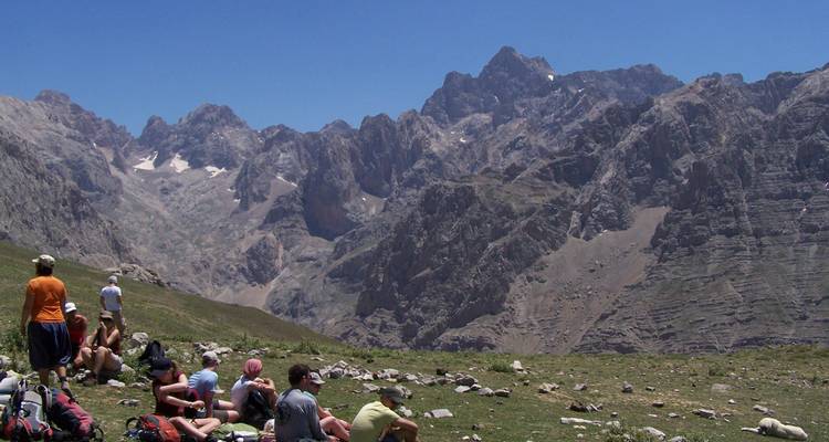 Un groupe de randonneurs se repose sur une prairie alpine herbeuse face à des pics déchiquetés spectaculaires sous un ciel dégagé.