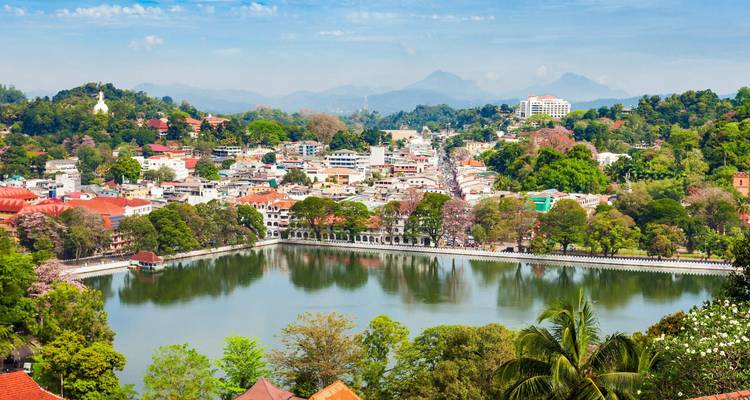 Panoramablick auf die Stadt Kandy, die einen spiegelnden See umschließt, mit Bergketten in der Ferne.