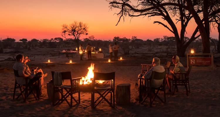 Reizigers zitten rond een gloeiend kampvuur onder een levendige oranje zonsonderganghemel in de bush