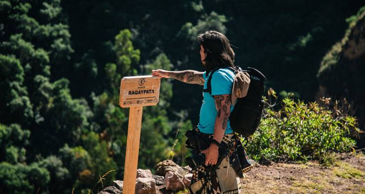Randonneur tatoué pointant vers un panneau de sentier en bois qui indique 'Raqaypata' sur fond de canyon luxuriant.