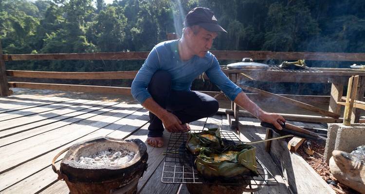 Local man grilling food on charcoal stove on wooden deck overlooking jungle