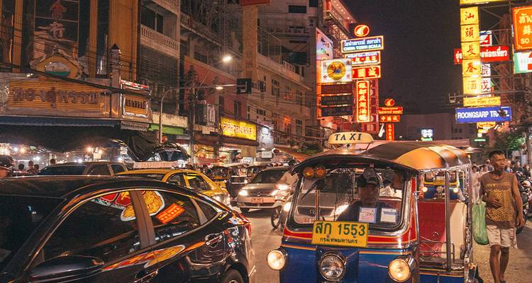 Bustling Bangkok street at night filled with neon signs, cars and a tuk-tuk