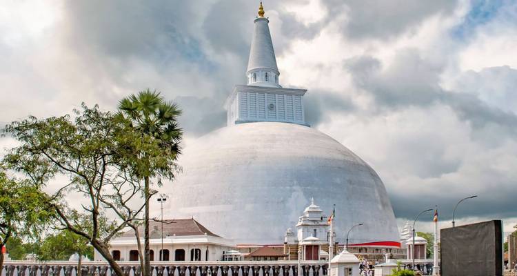 Massive weiße Kuppelstupa, die sich gegen einen dramatischen bewölkten Himmel erhebt, mit umliegenden Schreingebäuden.