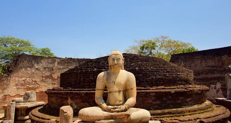 Antike sitzende Buddha-Statue vor kreisförmigen Backsteinruinen einer Stupa unter klarem blauen Himmel.