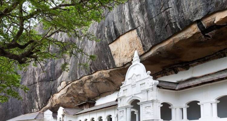 Weiße Tempelfassade, die unter einem überhängenden Felsenkliff zwischen grünen Bäumen errichtet wurde.