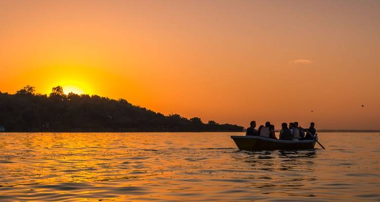 Silhouetted boat of passengers rowing across a lake at vivid orange sunset.