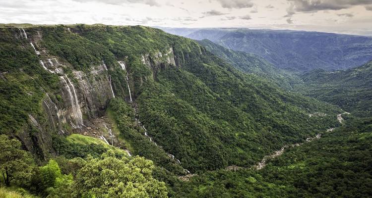 Vast green valley with multiple waterfalls cascading down sheer cliffs under broken clouds.