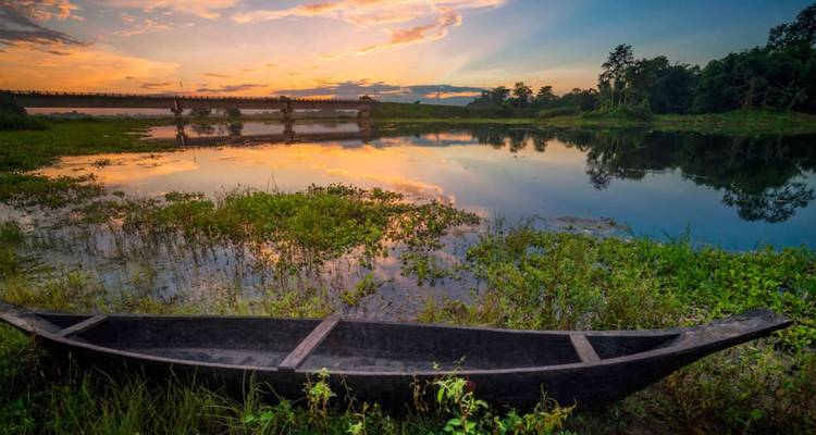 Wooden canoe beside a reflective river at colorful sunset with bridge in the distance.