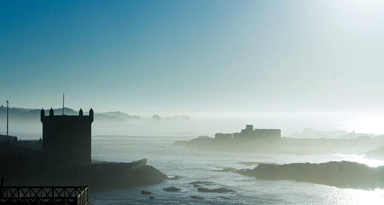 Misty coastal scene with silhouettes of fortifications and rocky shoreline at dawn.