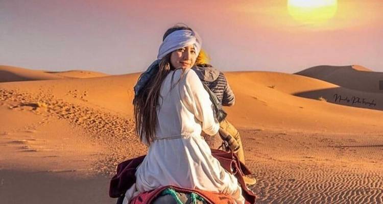 Una mujer vestida de blanco flotante monta un camello hacia el sol poniente sobre las dunas.