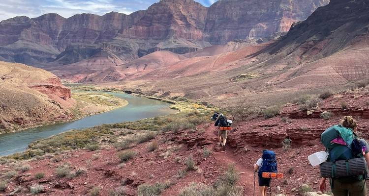 Rugzaktoeristen volgen een pad boven een slingerende rivier in een roodrotsen canyon.