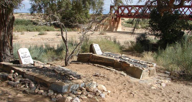 Oude graven overschaduwd door een eenzame boom staan naast een rode ijzeren brug in de woestijn.