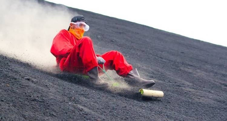 Traveler sandboarding down dark ash slope in red suit and goggles raising dust plume.