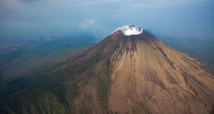 Aerial view of steep stratovolcano with steaming crater and radial lava flows through forest.