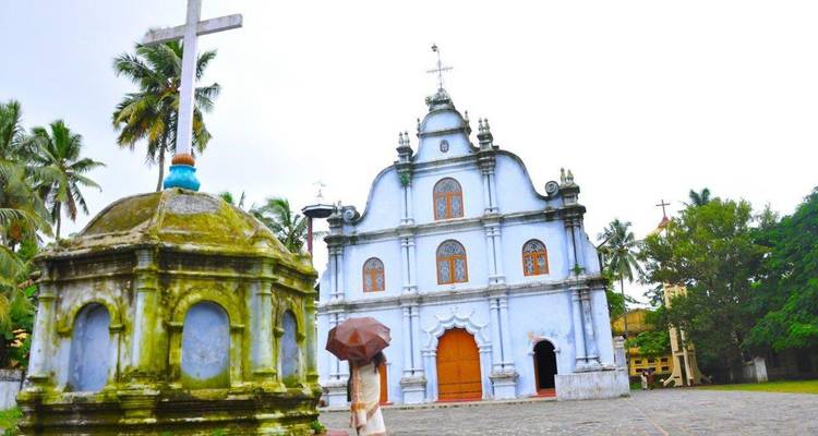 Bezoeker met een paraplu loopt naar een historische witgekalkte koloniale kerk omringd door palmbomen.
