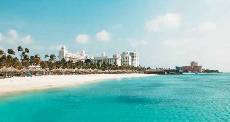 Wide view of a white-sand Caribbean beach with turquoise water and a grand beachfront resort lined with palm trees under a bright blue sky