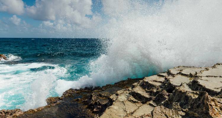 Powerful ocean waves crash against jagged coastal rocks sending white spray high into the air beneath scattered clouds