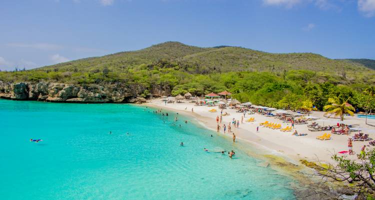 A curved Caribbean cove with turquoise water, white sand, umbrellas and swimmers set against low green hills