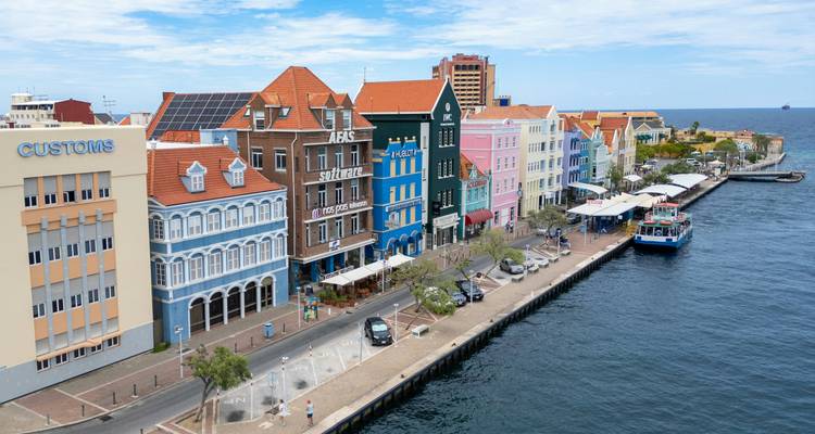 Colorful Dutch-colonial buildings line the waterfront promenade beside a deep blue harbor under a partly cloudy sky