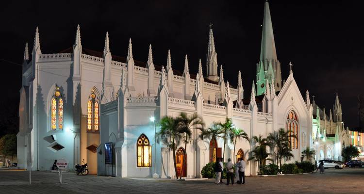 Église néo-gothique blanche illuminée avec vitraux et hautes flèches la nuit, avec un petit groupe qui discute à l'extérieur.