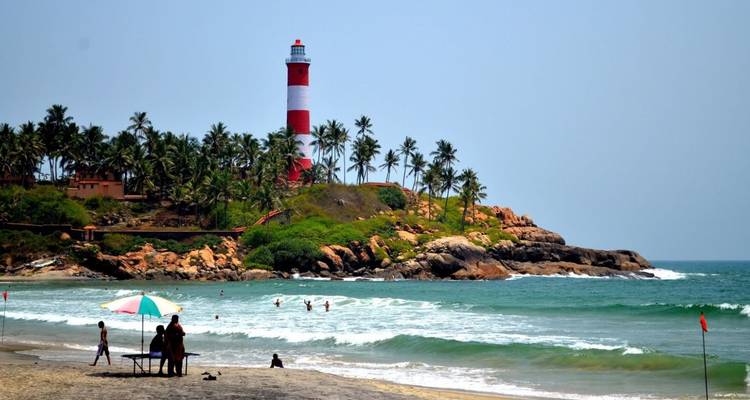 Red-and-white striped lighthouse on a rocky palm-covered promontory overlooking swimmers and beachgoers at Kovalam beach.