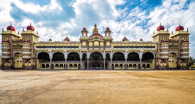 Der majestätische Mysore-Palast mit roten Kuppeln und verzierten Bögen beherrscht einen weitläufigen Innenhof unter teilweise bewölktem Himmel.
