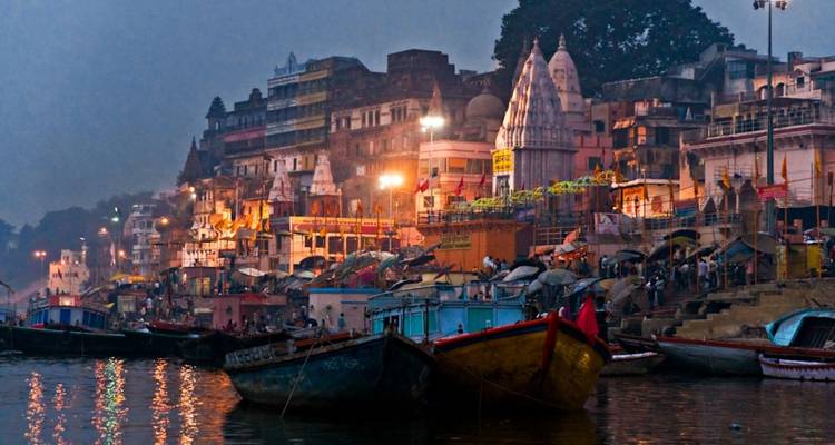 Verlichte ghats van Varanasi bij schemering met gelaagde gebouwen aan de rivier en kleurrijke boten langs de Ganges.
